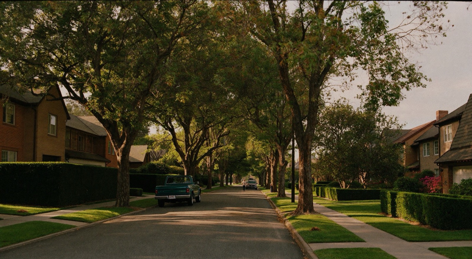 A tree-lined residential street at golden hour, with brick houses, trimmed hedges and a pickup parked at the curb.